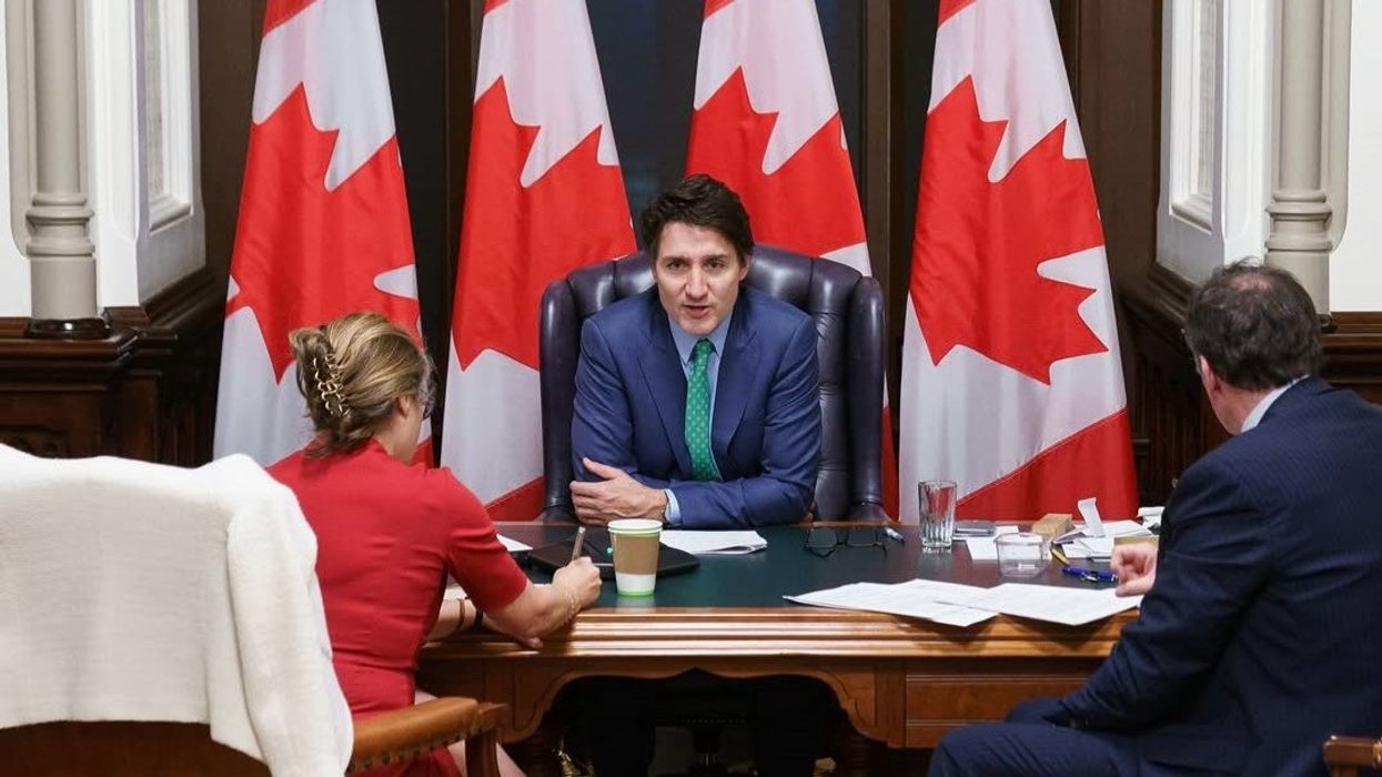 justin trudeau sitting at a desk in front of canadian flags in prime minister's office