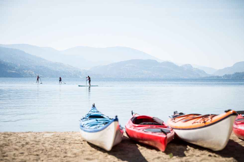 Kayaks on the sand of Okanagan Lake while people paddle board in the water.