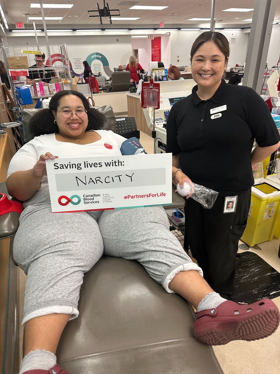 Kianni Reynolds-Lewis in a chair at the donor centre holding a sign that reads "Saving lives with: Narcity." Registered nurse Percy stands to the right, also smiling. 