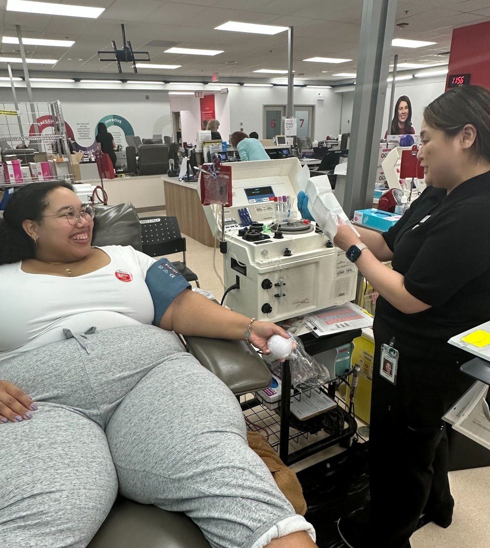 Kianni Reynolds-Lewis in a chair at the donor centre, smililing at registered nurse Percy who is preparing the apheresis machine for the plasma donation. 