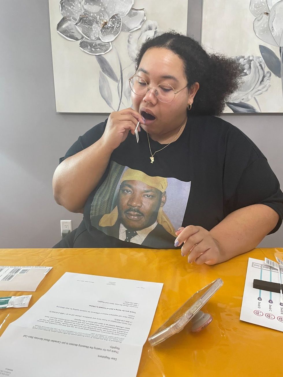 Kianni Reynolds-Lewis sits at a table strewn with information about the stem cell donation process, swabbing the inside of her cheel with a buccal swab. 