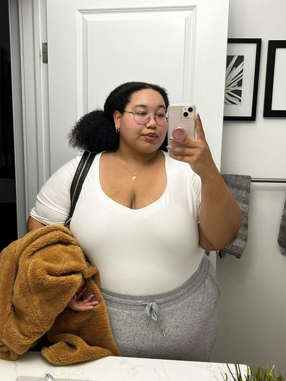 Kianni Reynolds-Lewis takes a selfie in a mirror while holding a fuzzy brown jacket and wearing a white tee and grey sweatpants. 