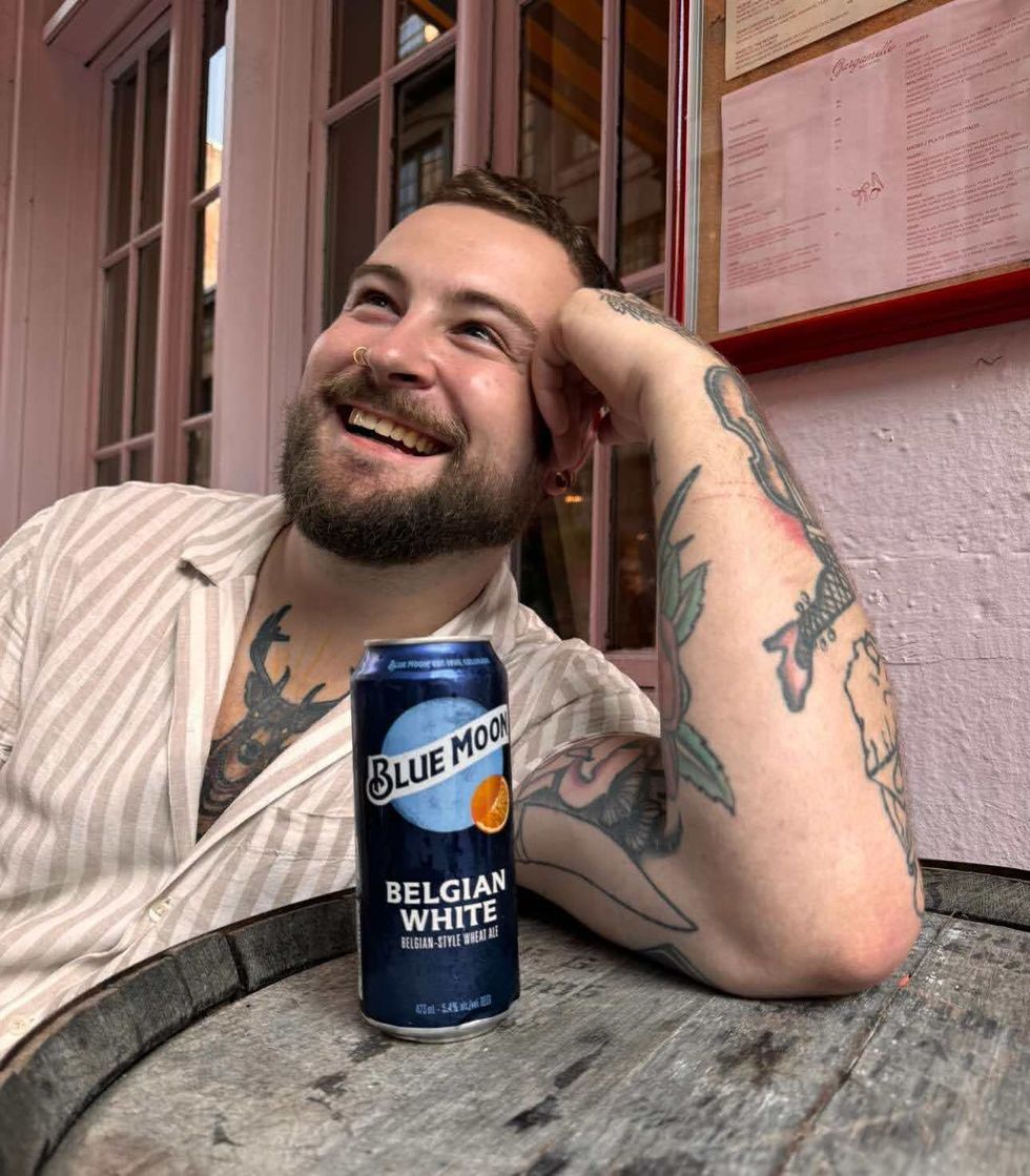 Lo\u00efc Fortin sourit en reposant sa t\u00eate sur sa main et son coude sur un baril avec une canette de Blue Moon Belgian White \u00e0 c\u00f4t\u00e9 de lui.