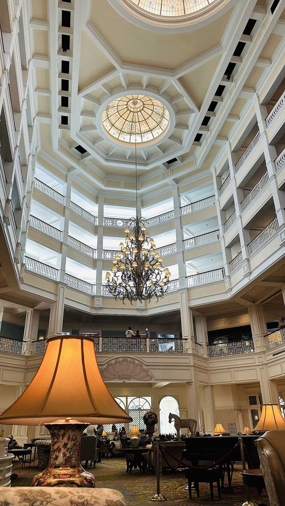 Looking up at the five-storey open atrium of Disney's Grand Floridian Resort lobby, featuring a large ornate chandelier, stained glass dome skylight, wrought iron balcony railings on each level, and warm lamp light in the foreground.