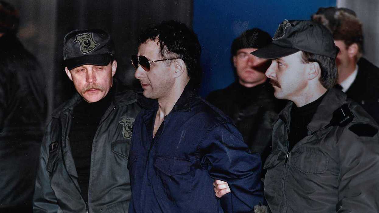 Man in a prison uniform with hands bound behind his back being escorted by police officers.