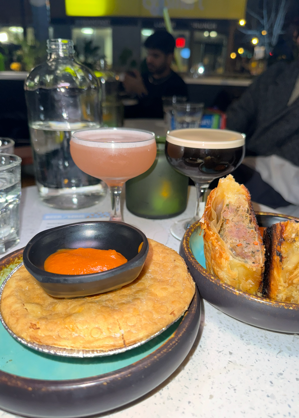 Meat pie with tomato sauce, beef sausage rolls and two cocktails on a table.