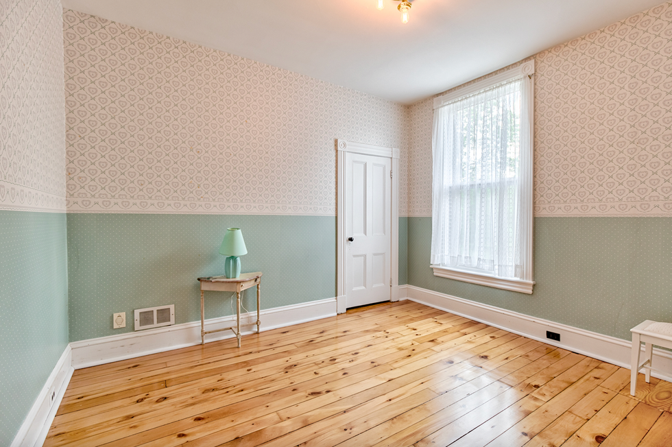 One of the bedrooms in the mansion that has green and patterned wall paper, a window with a see-through curtain and light-coloured flooring.