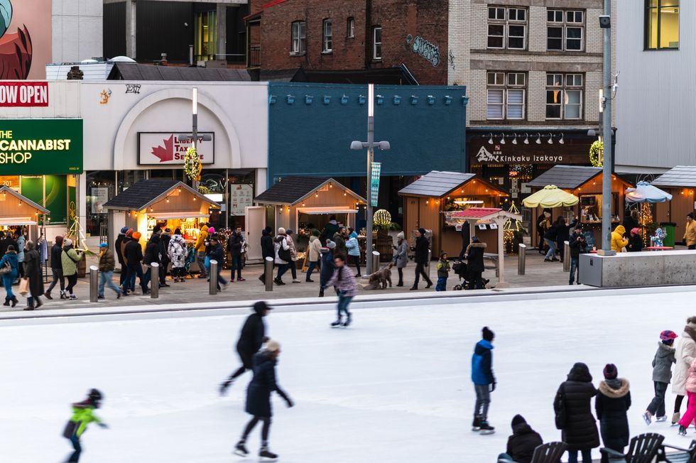 People are seen skating on an outdoor rink surrounded by glowing wooden kiosks.