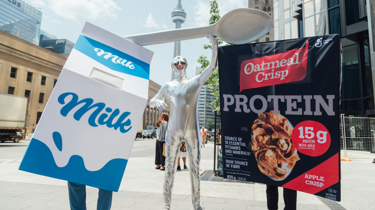 People dressed as a milk carton, oatmeal cereal box, and silver spoon costume pose on a city street with the CN Tower in the background during a promotional event.