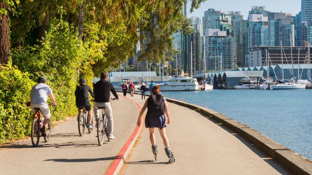 People on bikes and roller blades on a path next to trees on the left side and the ocean on the right side with tall buildings and boats nearby.