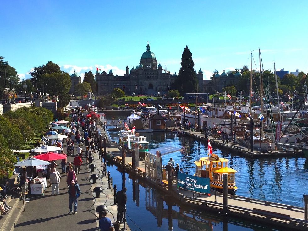 People walking along the water that is filled with boats and a parliament building in the background.