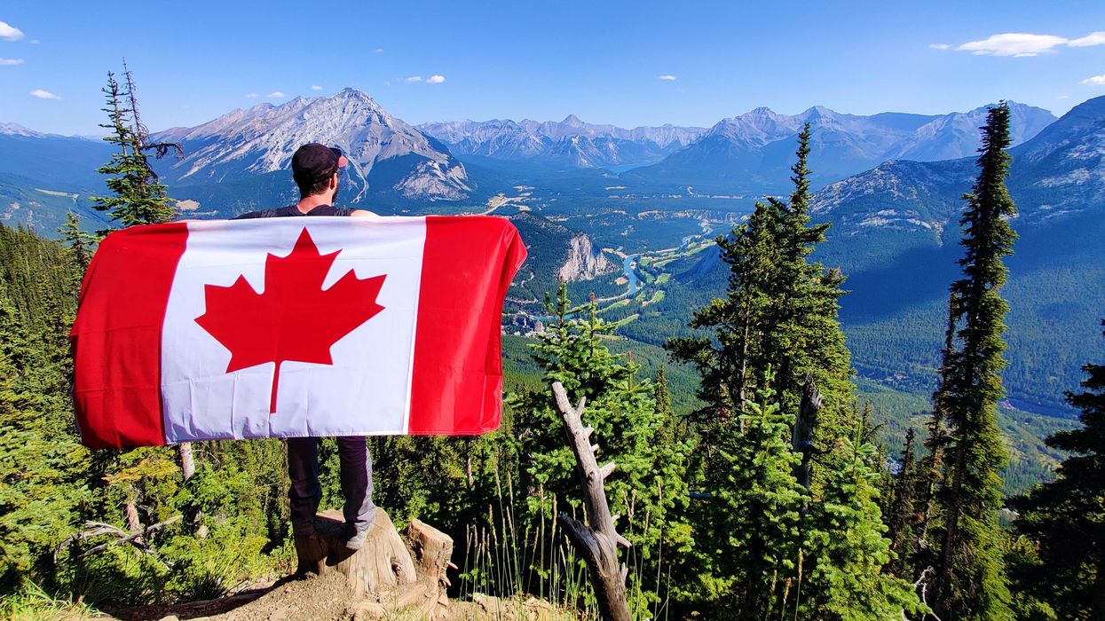 Person holding a Canadian flag on the edge of a cliff overlooking a mountain range.