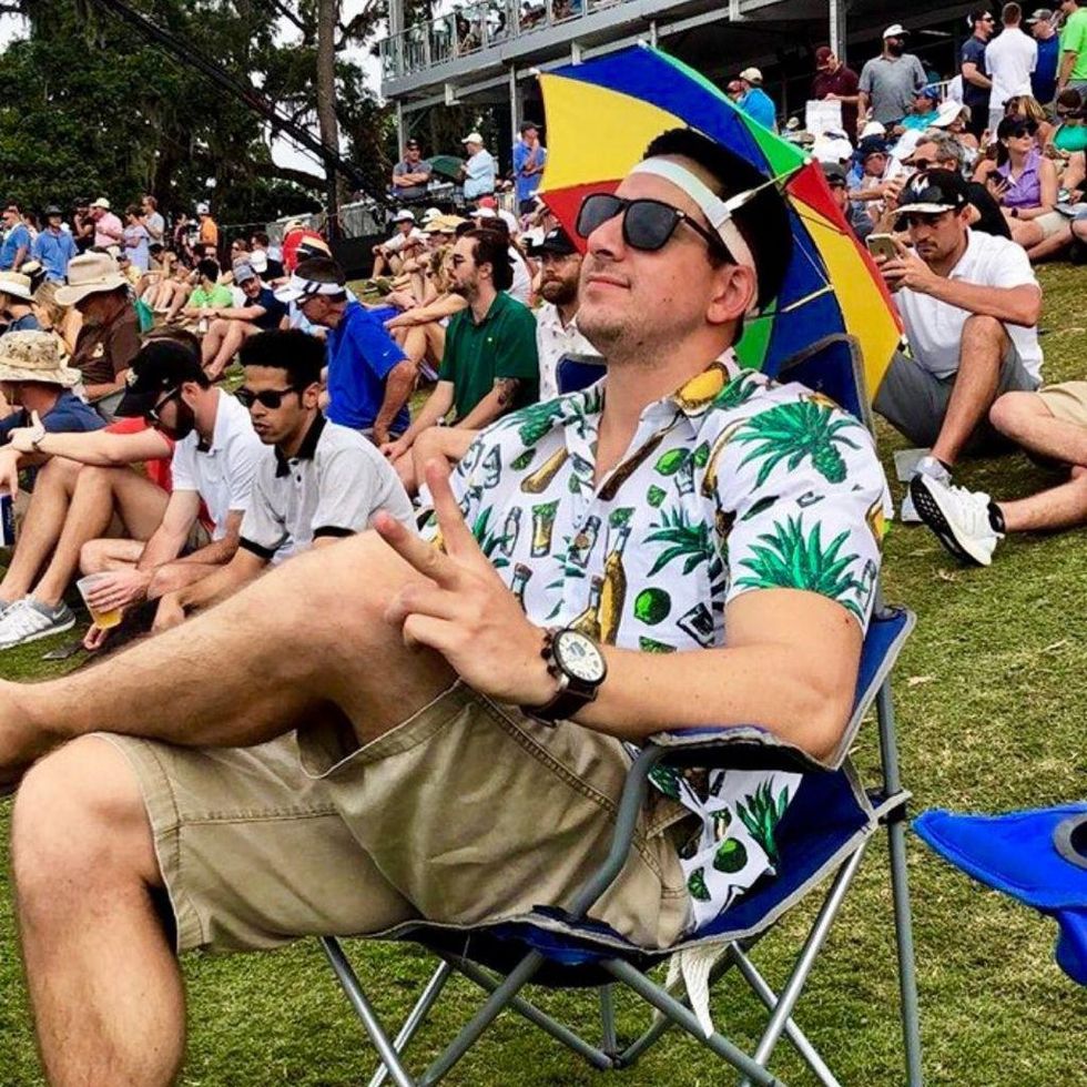 person sitting in a foldable chair in a park full of people with the tequila Hawaiian shirt