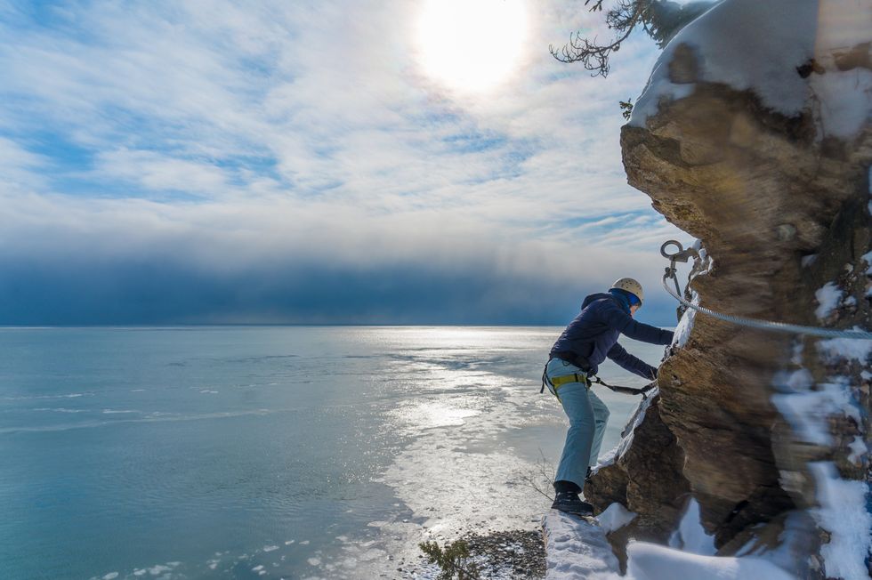 Personne d\u00e9ambulant le long des flancs enneig\u00e9s d'une montagne, utilisant des c\u00e2bles pour se d\u00e9placer, avec en arri\u00e8re-plan le panorama du fleuve.