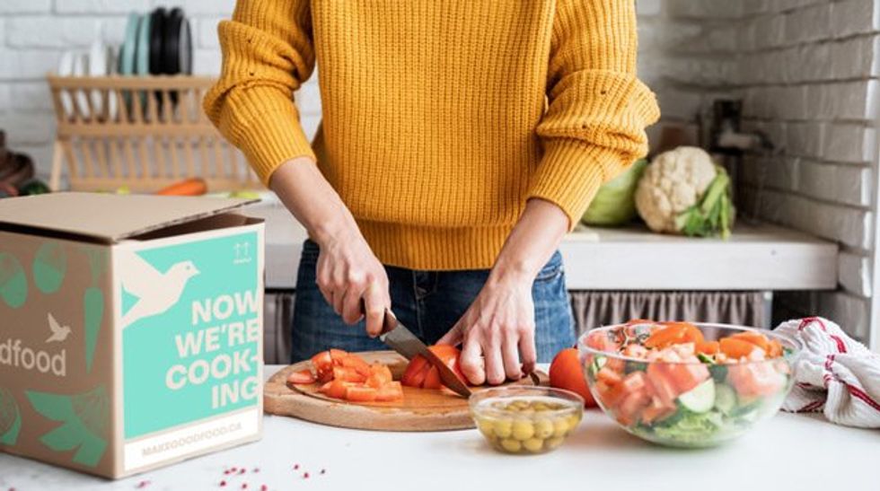 Personne en train de d\u00e9couper une tomate sur une planche entour\u00e9e d'ingr\u00e9dients pour la pr\u00e9paration d'un repas, avec une bo\u00eete Goodfood \u00e0 proximit\u00e9.