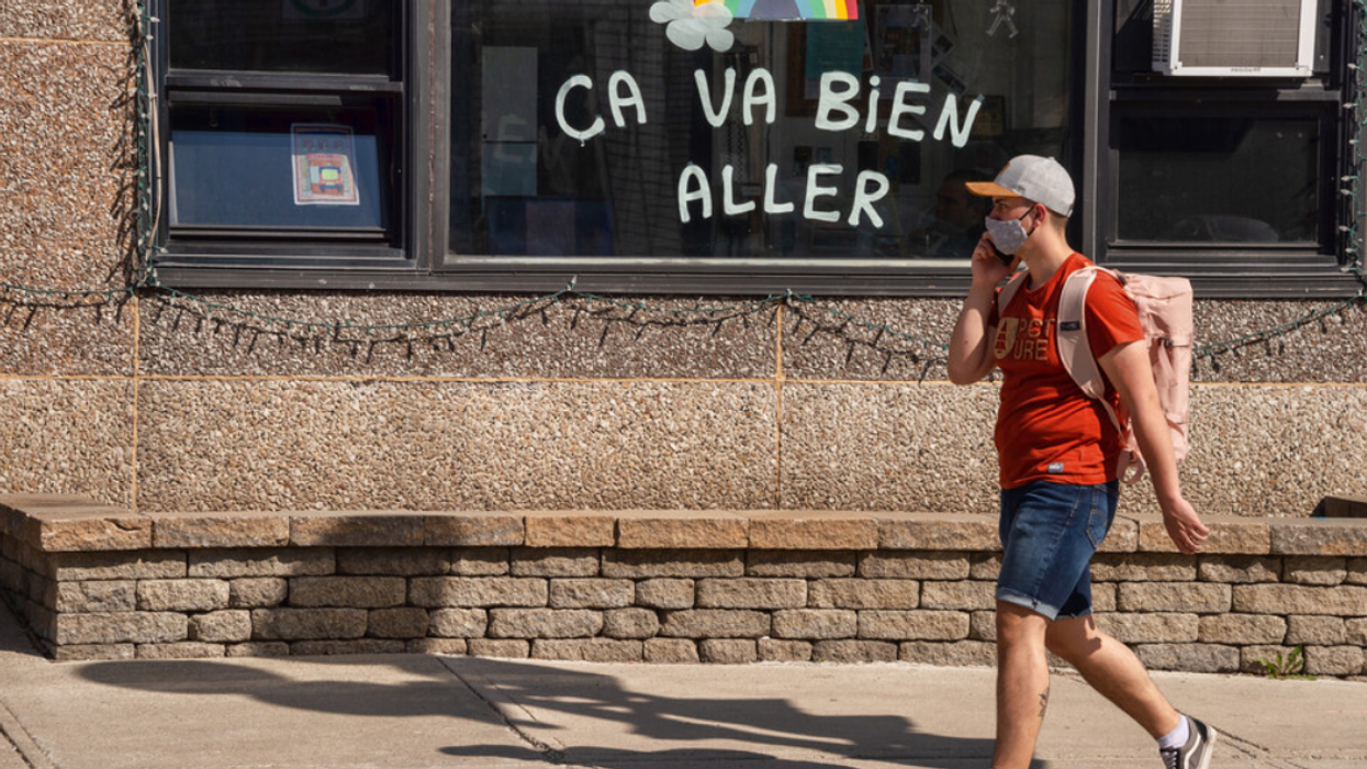 Personne masquée marche sur le trottoir devant une vitrine avec un arc-en-ciel et un « Ça va bien aller ».