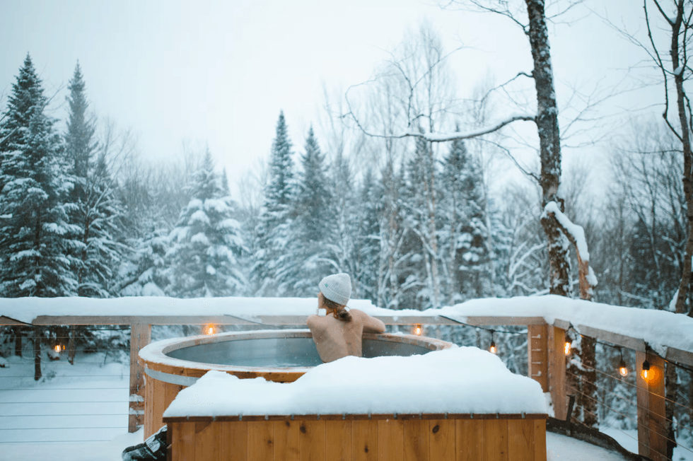 Personne portant une tuque, assise dans un spa de c\u00e8dre sur une terrasse enneig\u00e9e entour\u00e9e de sapins et de lumi\u00e8res suspendues.
