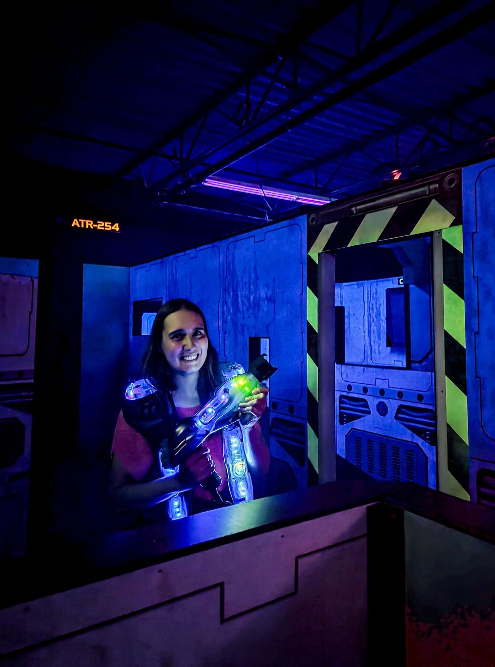 Personne souriante jouant au laser tag dans une ar\u00e8ne illumin\u00e9e de n\u00e9ons bleus et verts.