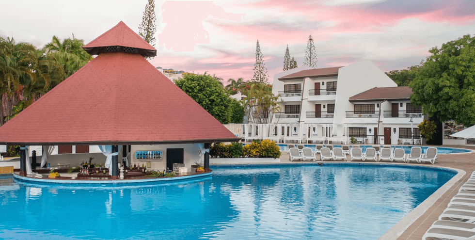 Piscine ext\u00e9rieure avec bar int\u00e9gr\u00e9, chaises longues et b\u00e2timents blancs entour\u00e9s de palmiers, au BlueBay Villas Doradas \u00e0 Puerto Plata.