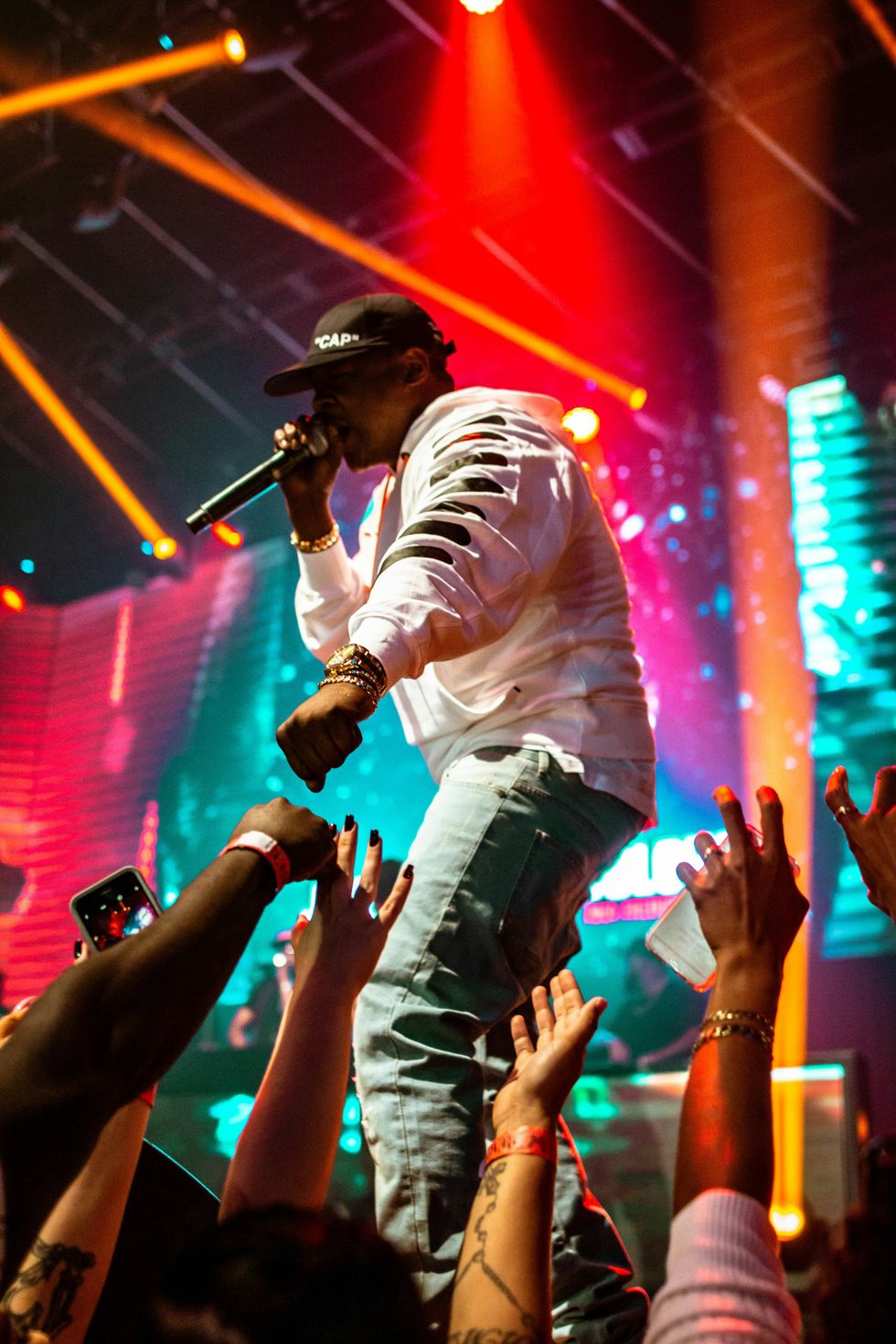 Rapper performing on stage in a white hoodie and jeans, holding a microphone while engaging with the crowd under colorful stage lights.