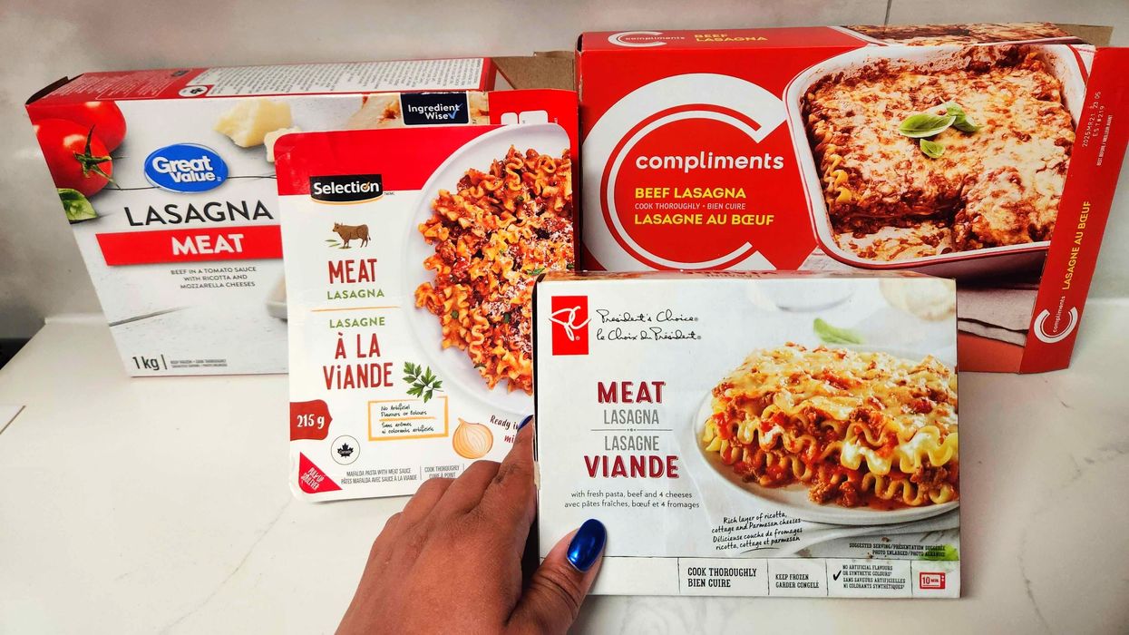 Red and white voxes of lasagna on a white marble counter top with one box being held by a hand with blue nails.