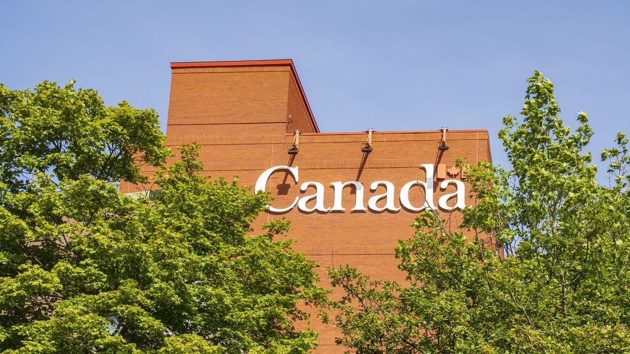 Red brick building with lettering reading "Canada," with trees in the foreground and a clear blue sky behind.