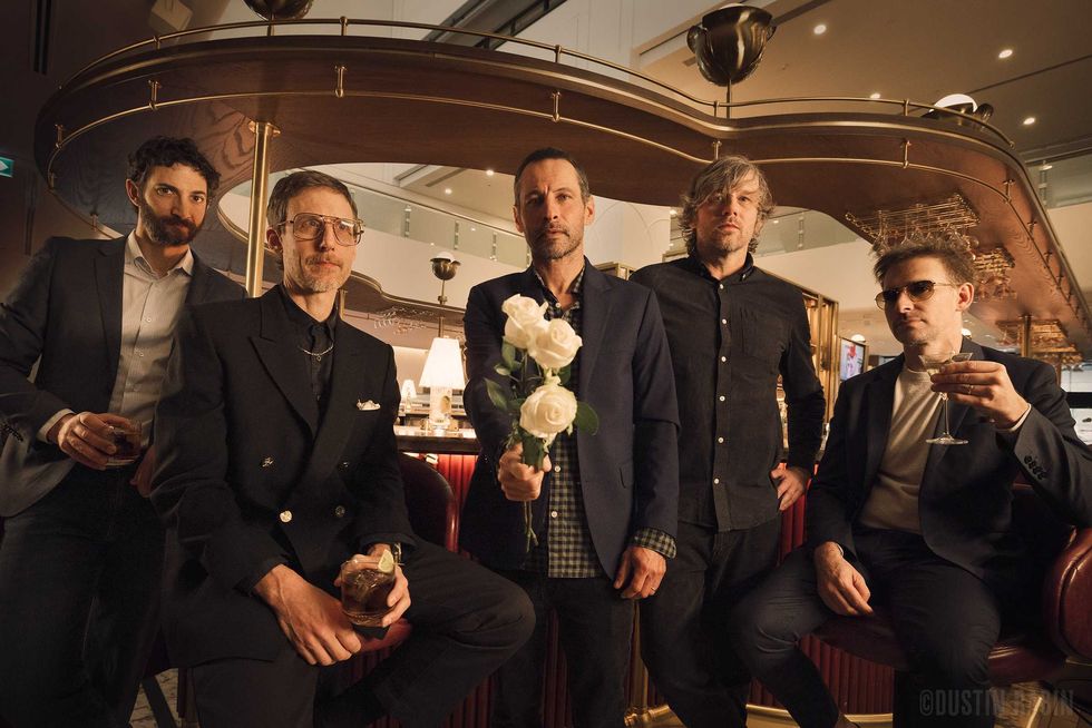 Sam Roberts Band in a bar wearing black suits, drinking cocktails. Sam Robets, centre, holds out three white roses.