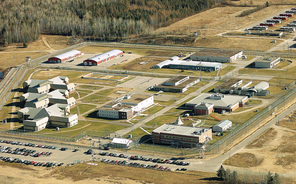Several buildings on a piece of land surrounded by a fence and a parking lot filled with cars.