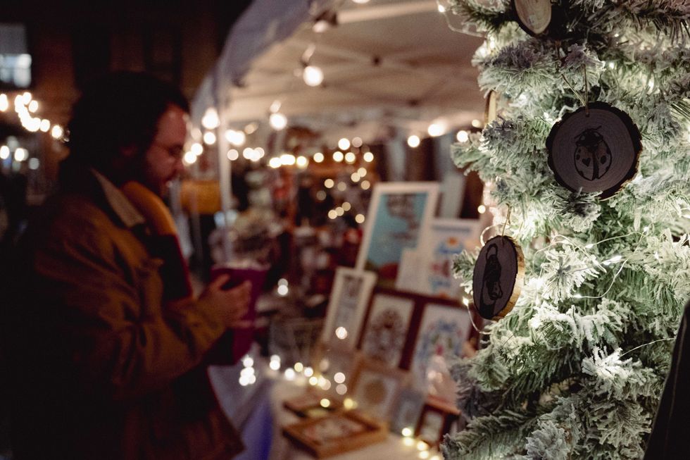 Shoppers browsing crafts at a night market stall lit by holiday lights.