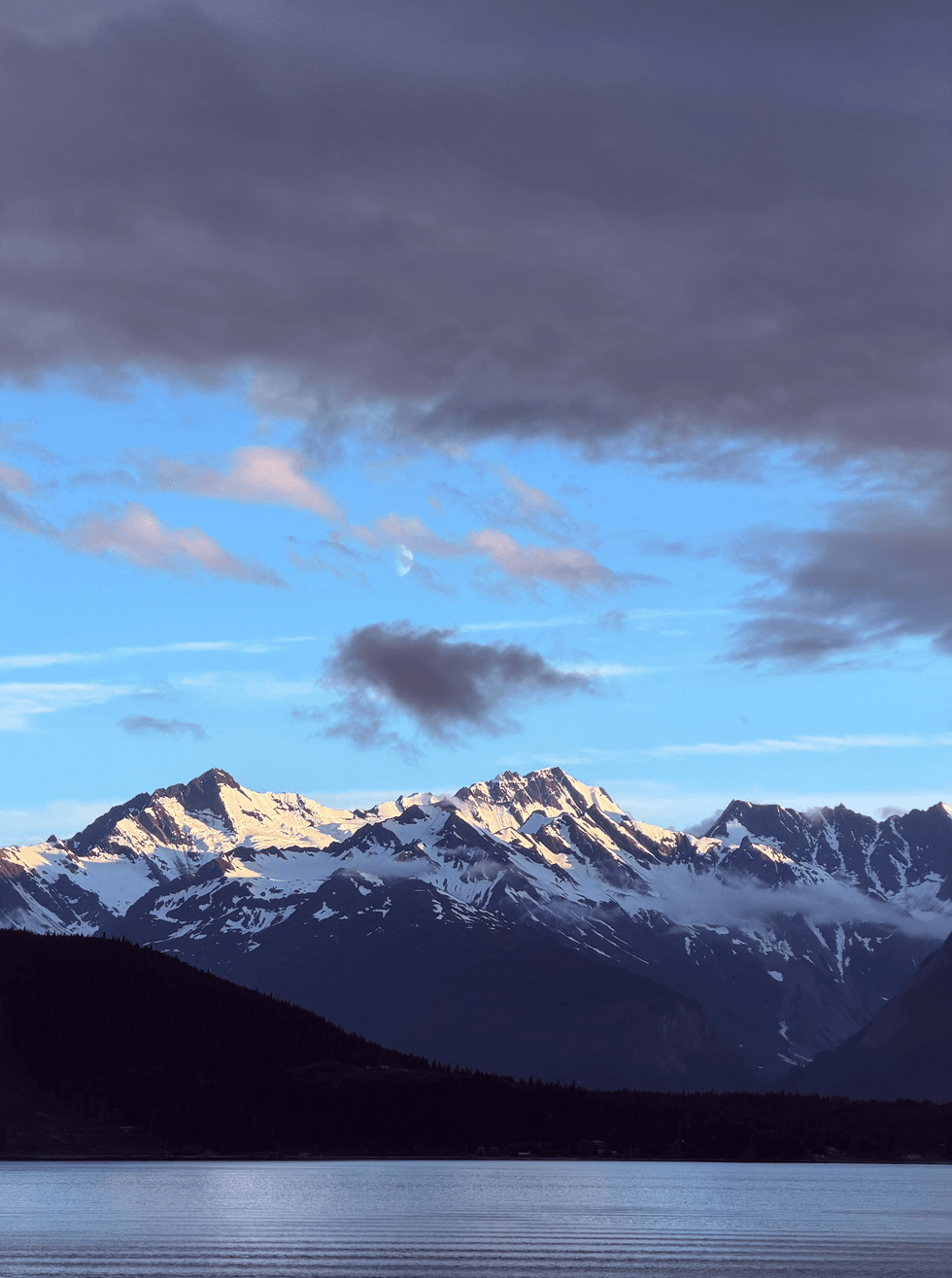Snow-capped Alaska mountain range under a blue sky with clouds and a visible moon, with calm water in the foreground.