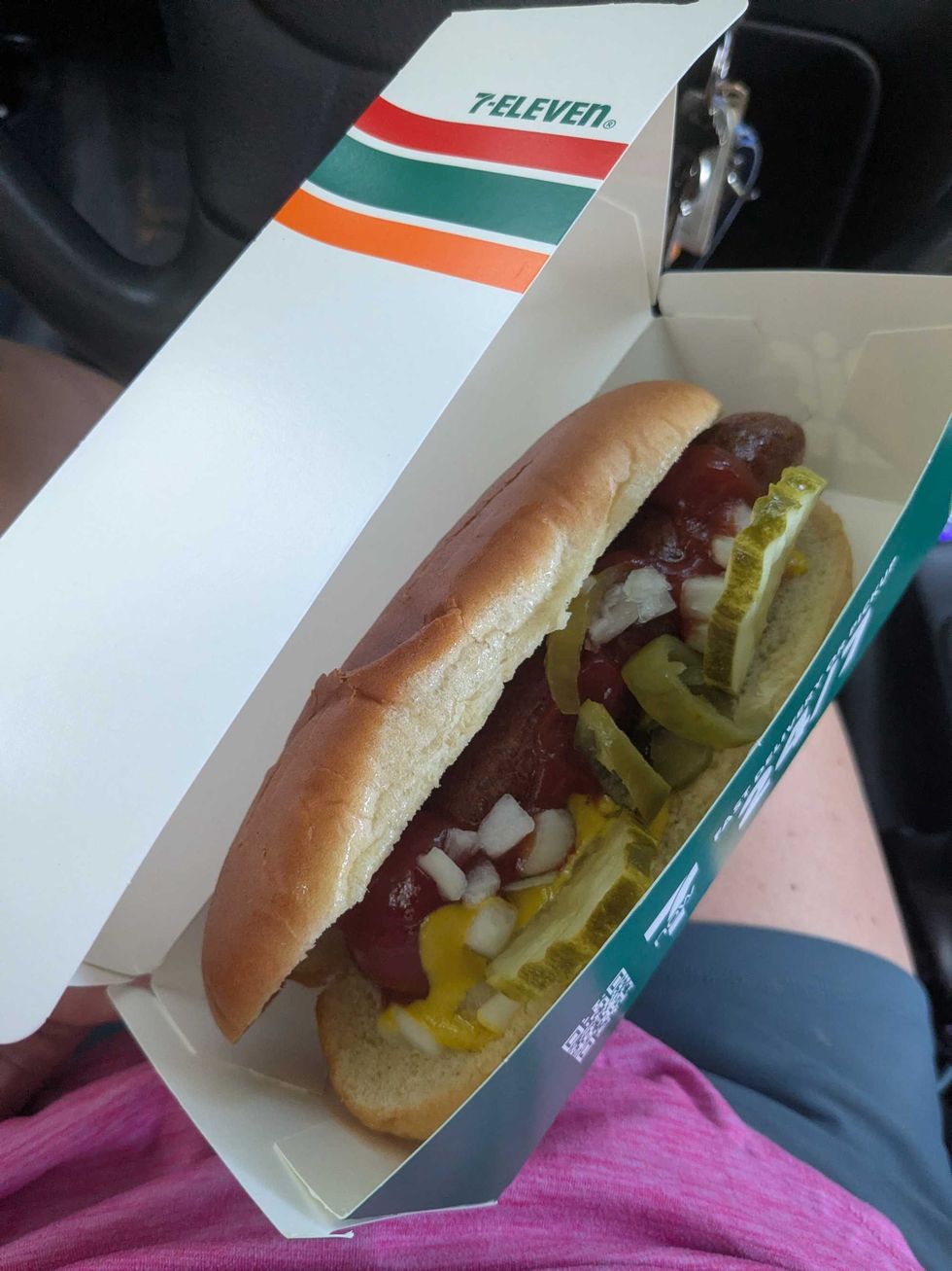 Someone holding a 7-11 hot dog in a cardboard box with lots of toppings on it
