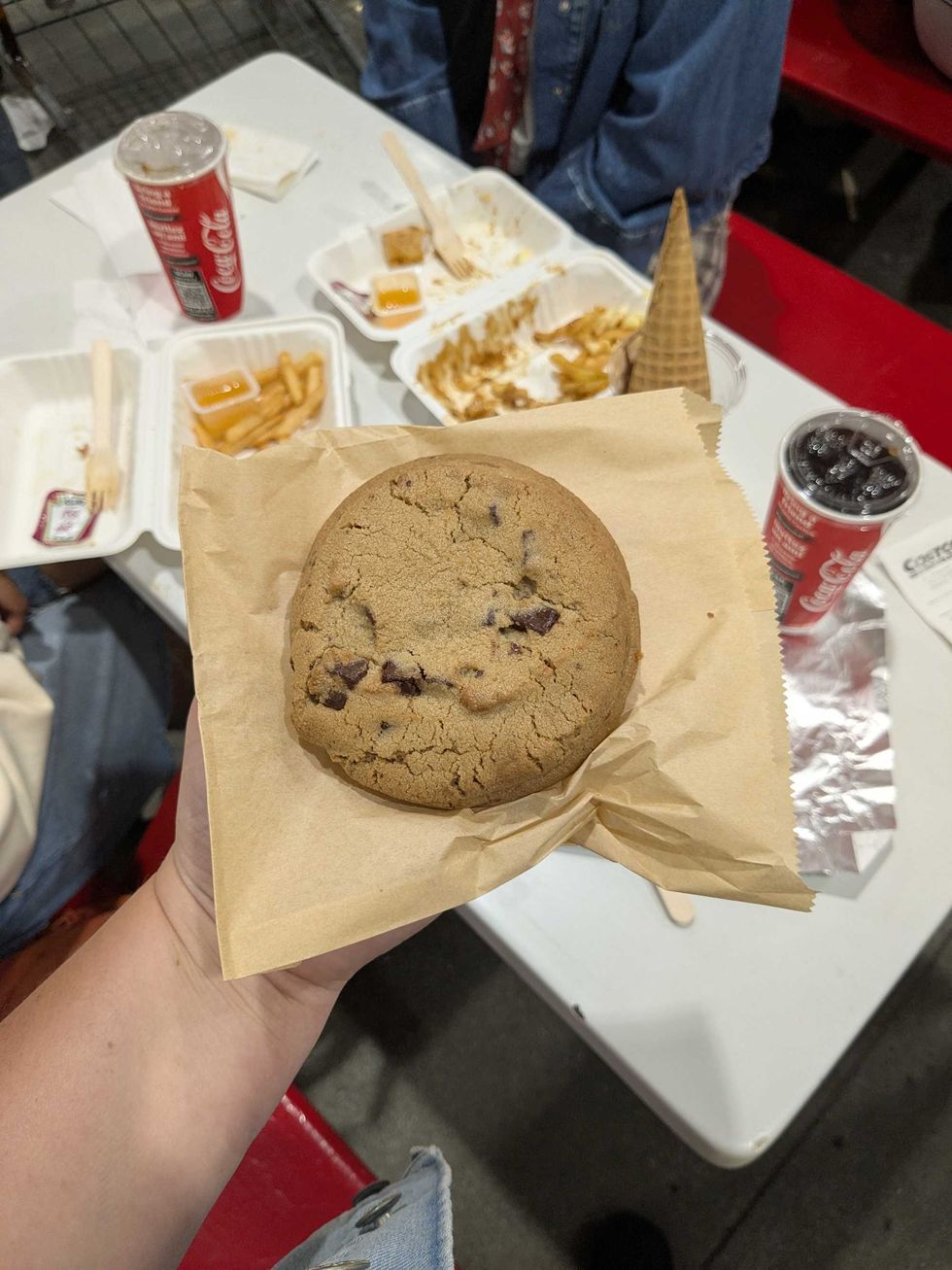 Someone holding a big chocolate chip cookie in front of a Costco table of food
