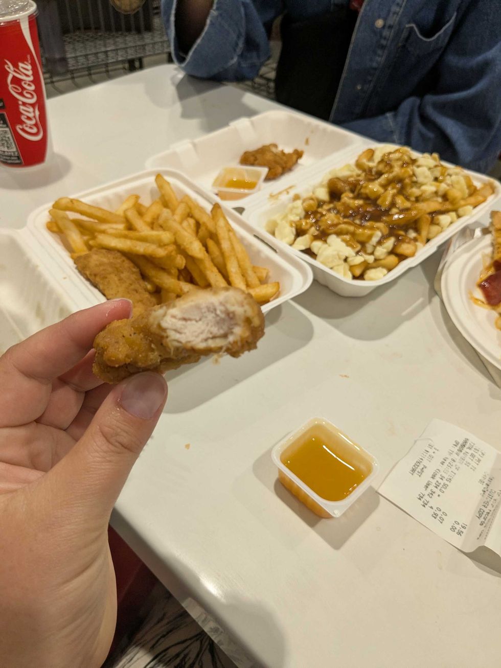 Someone holding a piece of a chicken strip in front of a table with other Costco food on it