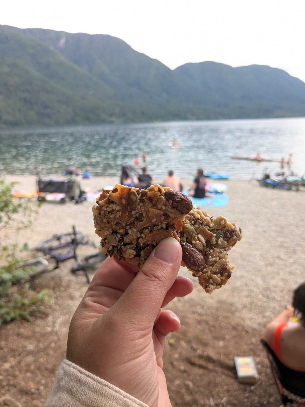 Someone holding pieces of nut and seed brittle up in front of a lake
