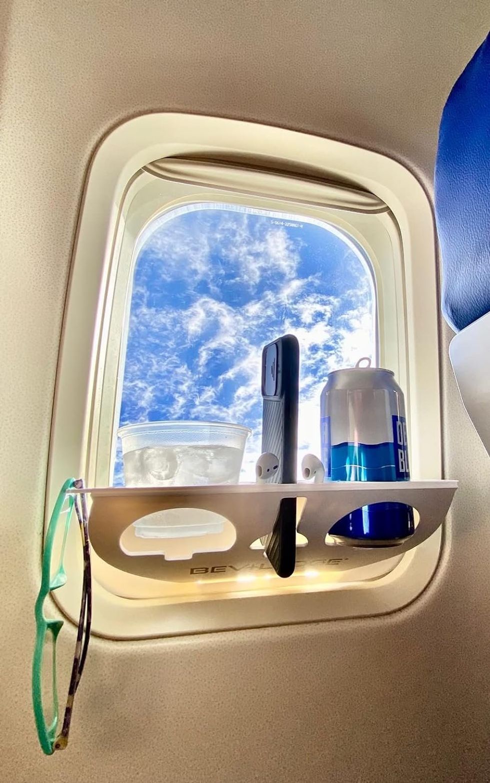 Station d'organisation de fen\u00eatre d'avion avec un verre, une cannette, un t\u00e9l\u00e9phone, des \u00e9couteurs et des lunettes.