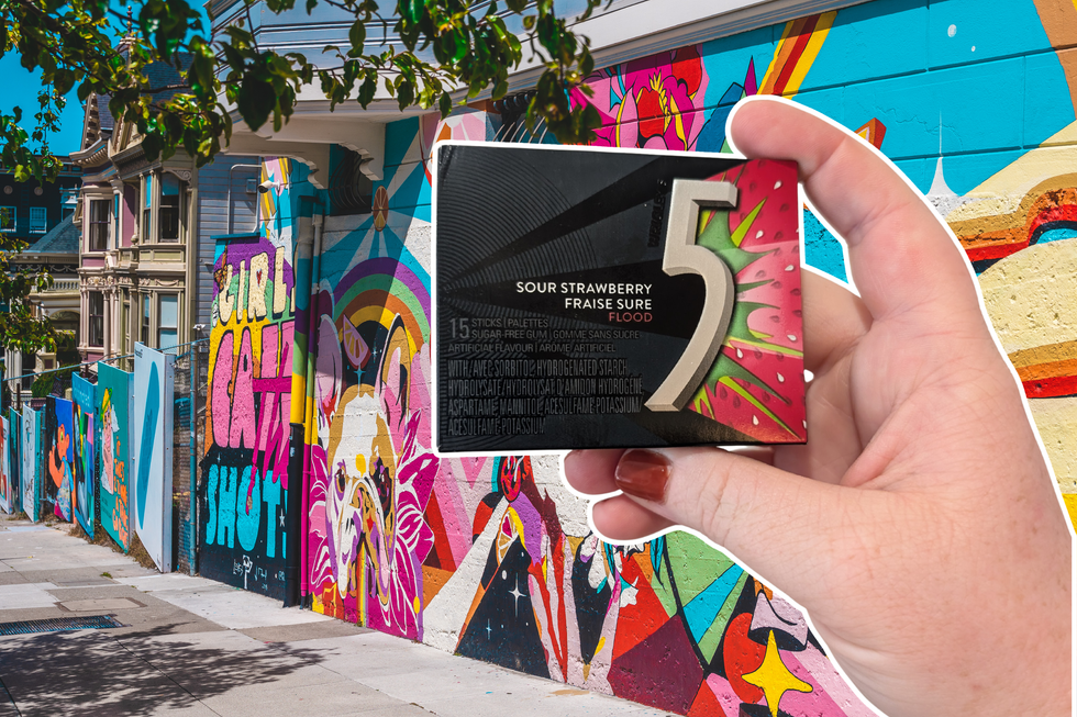 Tall fences on a city street are decorated with bright street art. A hand holding Flood flavour 5 Gum is on top.
