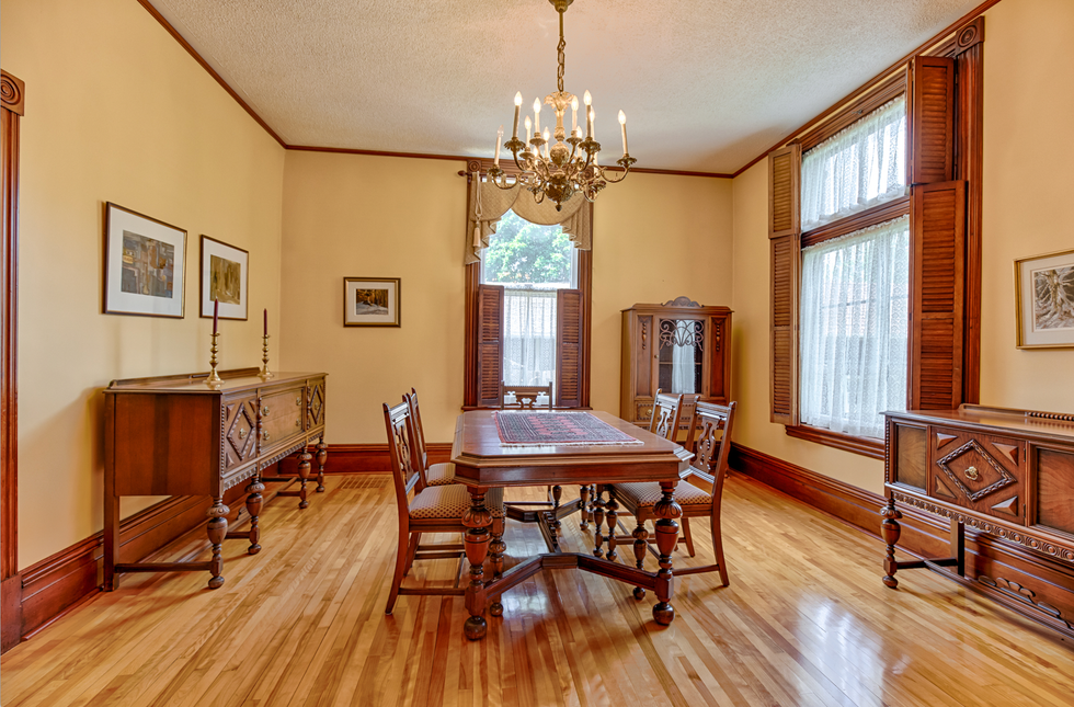 The a large wooden dining table with four chairs in a room, with a chandelier, large windows and wooden cabinets.
