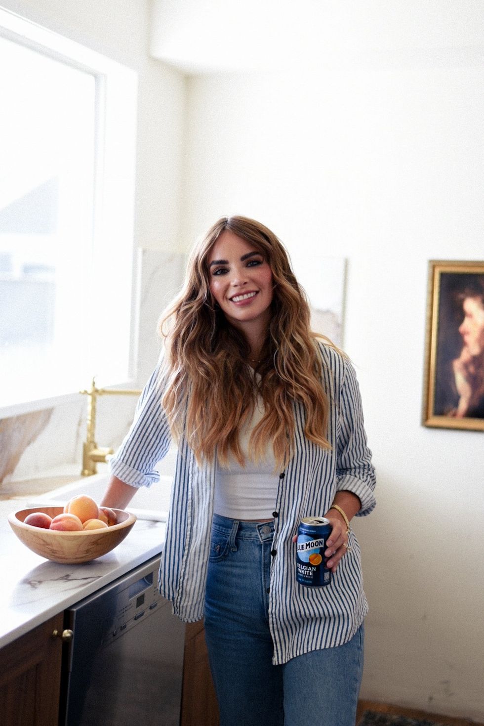 Thea VanHerwaarden stands in a kitchen wearing a blue striped shirt and jeans, holding a can of Blue Moon Belgian White.