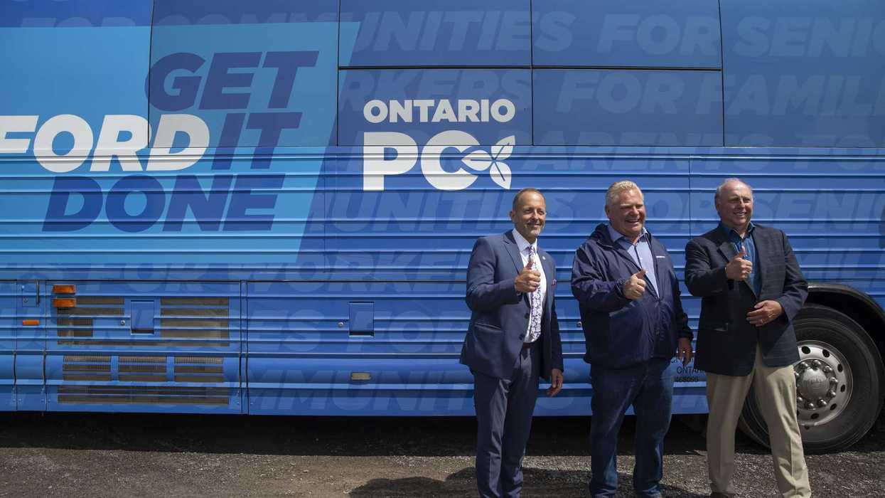 Three men give a thumbs-up posing in front of a Doug Ford campaign bus.