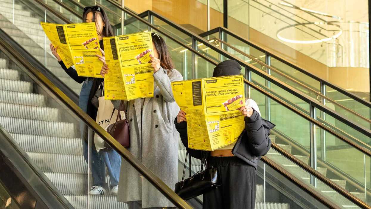 Three people ride an escalator while reading bright yellow No Name newspapers.