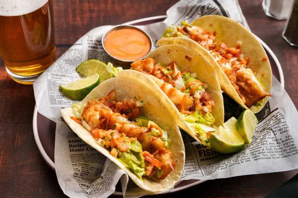 Three tacos on a newspaper-covered plate in a bistro, garnished with sauce and lime wedges.