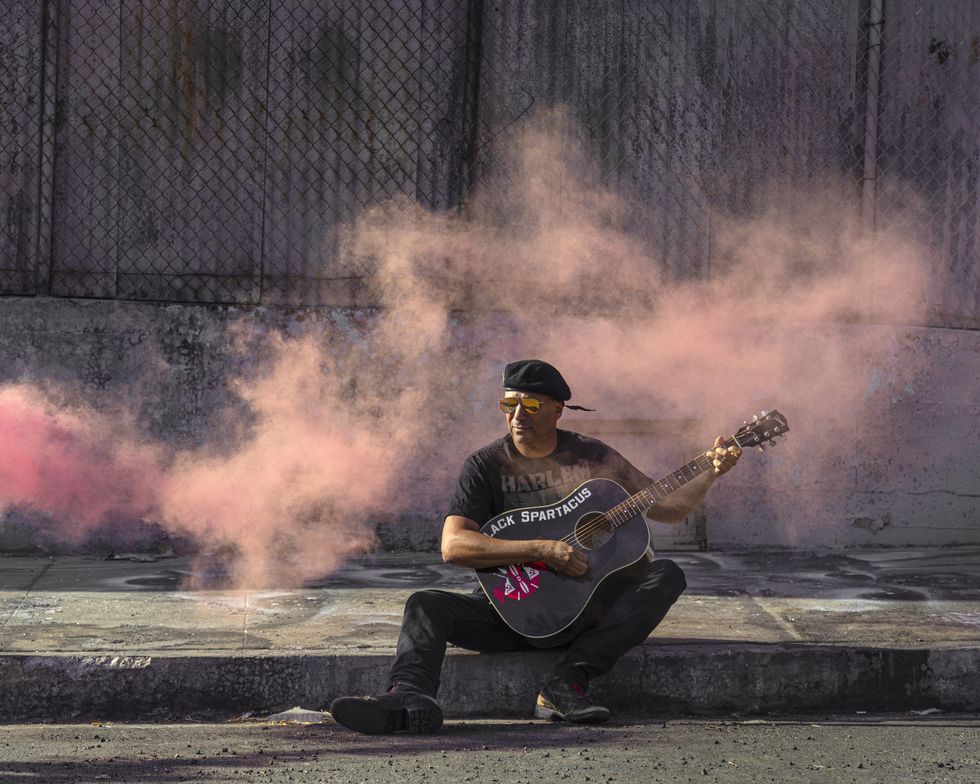 Tom Morello playing guitar on a curb while wearing a black beret with pink smoke drifting behind him. 