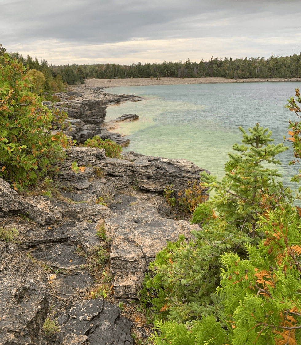 Turquoise-green water along a rugged limestone shoreline surrounded by evergreen trees and colourful fall foliage on Ontario's Bruce Peninsula.