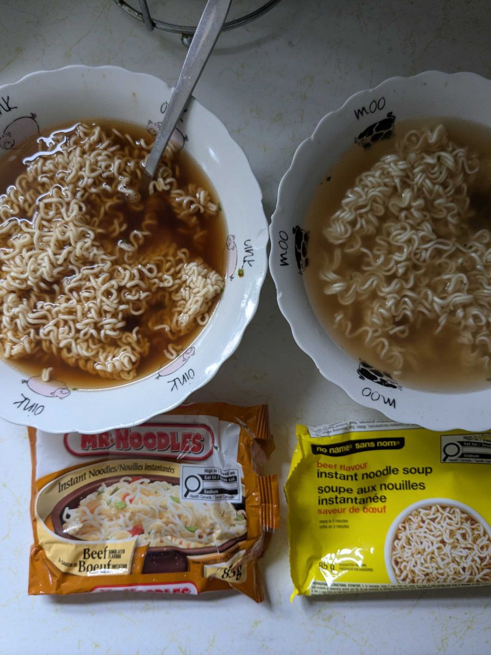 Two bowls of instant noodles side-by-side with the Mr Noodles and No Name packages beside them