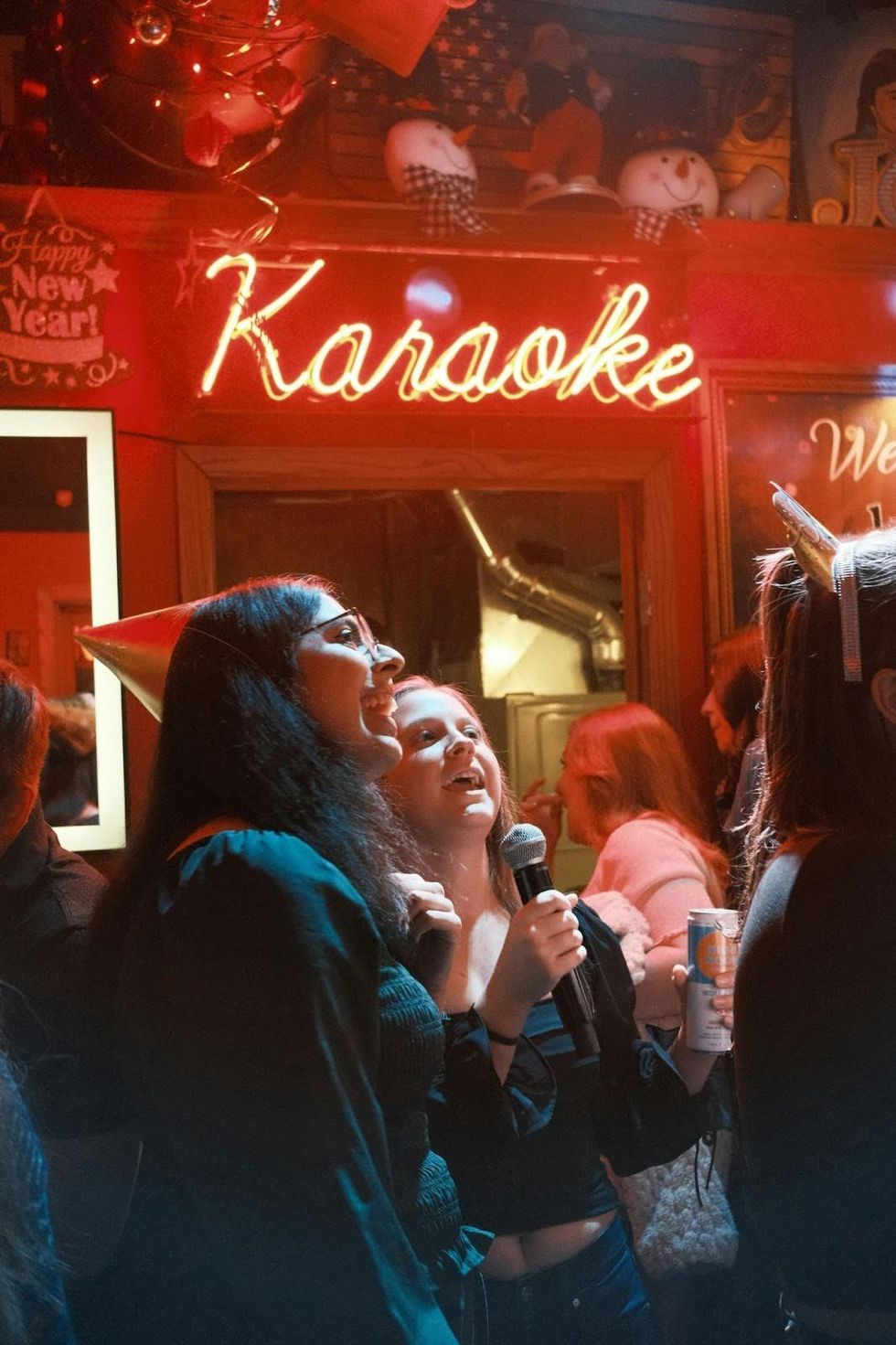 Two friends sing into a microphone. Behind them is a neon sign reading "karaoke."