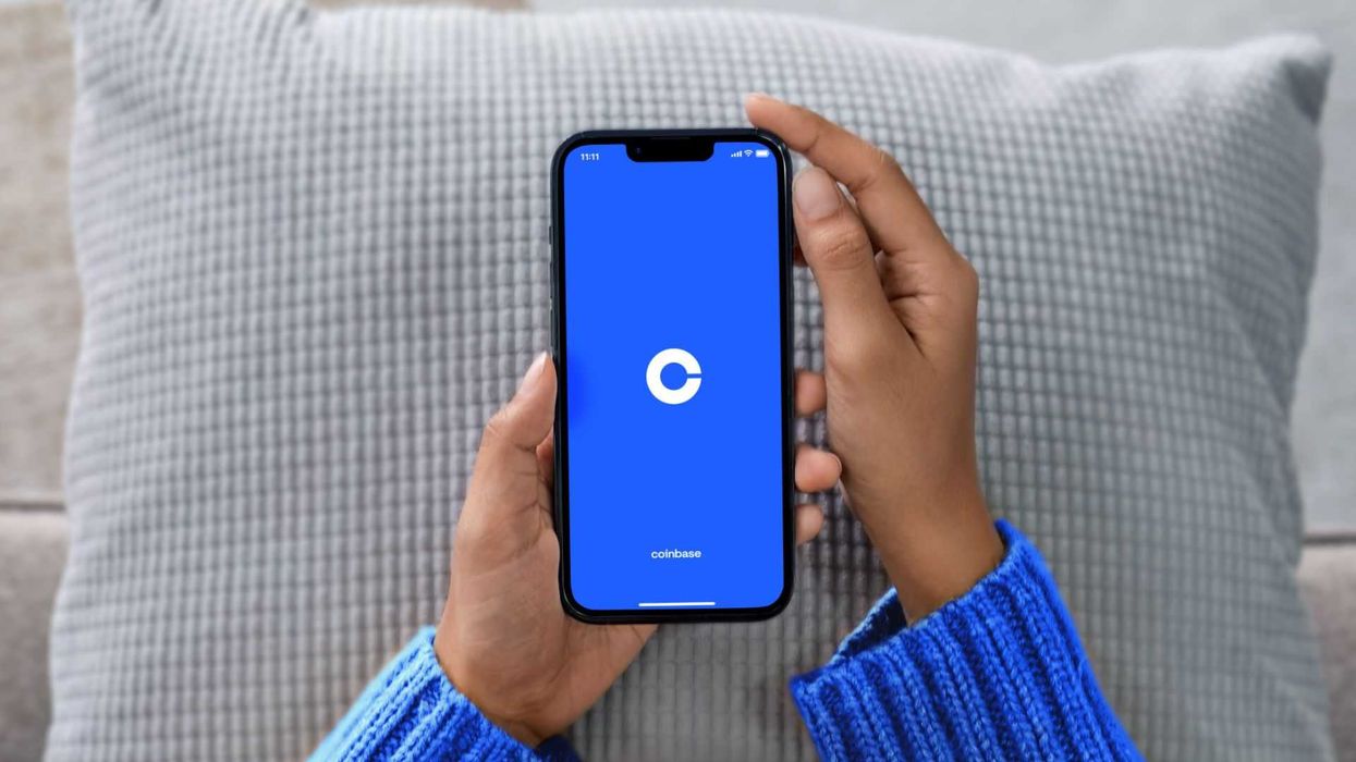 Two hands hold a phone, the screen of which displays the blue and white Coinbase logo, on top of a grey cushion.