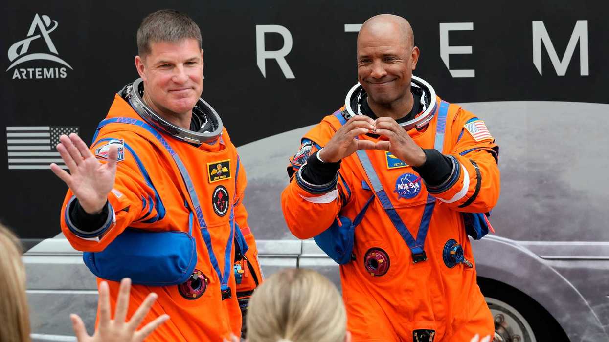 Two men in orange space suits wave at a crowd in front of a bus branded "Artemis."