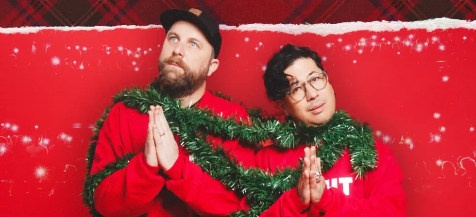 Two men in red sweaters pose against a festive red plaid-and-snow backdrop, wrapped together in green tinsel garland