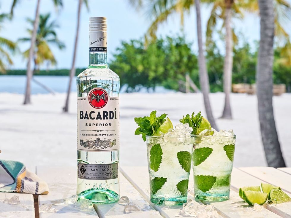 Two mojitos on a table with a bottle of BACARD\u00cd Rum in front of a beach with palm trees. 