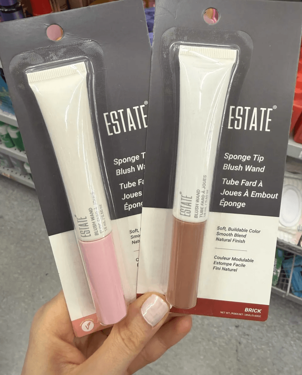 Two packaged Estate sponge tip blush wands in pink and brown held in a store aisle.